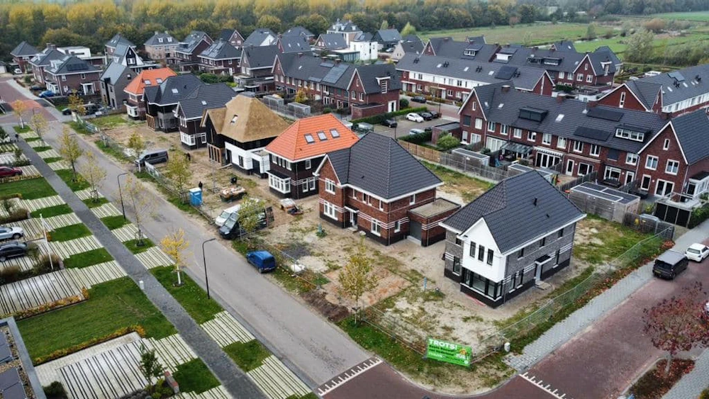 Luchtfoto van een nieuwbouwwijk in aanbouw met rijtjes bakstenen huizen, grijze en oranje daken, bouwmaterialen, auto's en groenstroken.