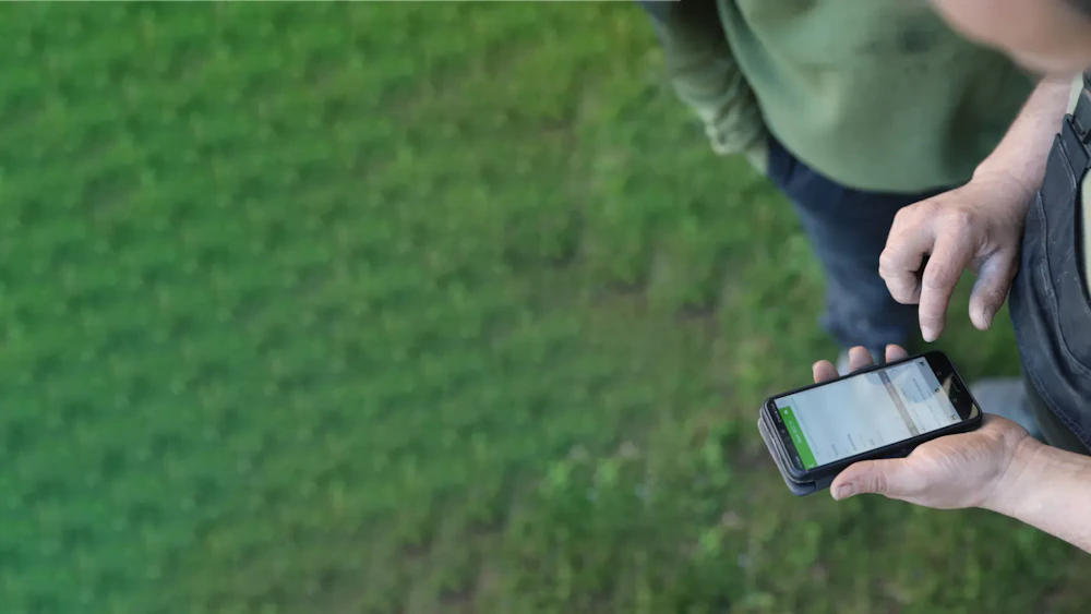 Een persoon houdt een smartphone vast bij een groen grasveld; het scherm toont een app.