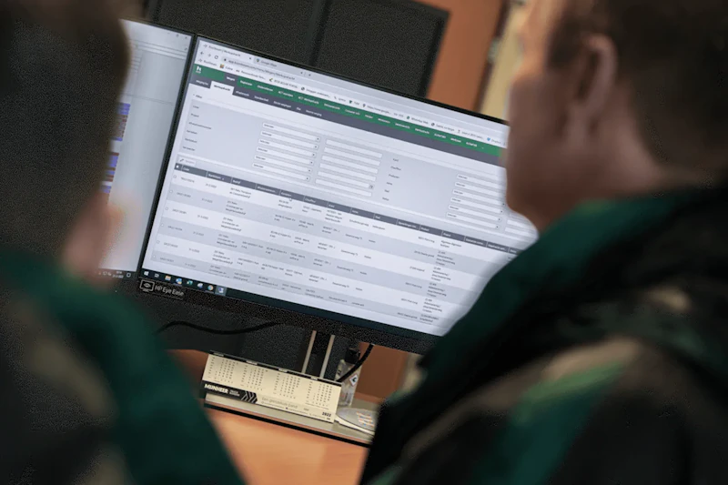 Twee personen kijken naar een monitor waarop een complexe webapp te zien is met formulieren en tabellen, en een groene navigatiebalk bovenaan.