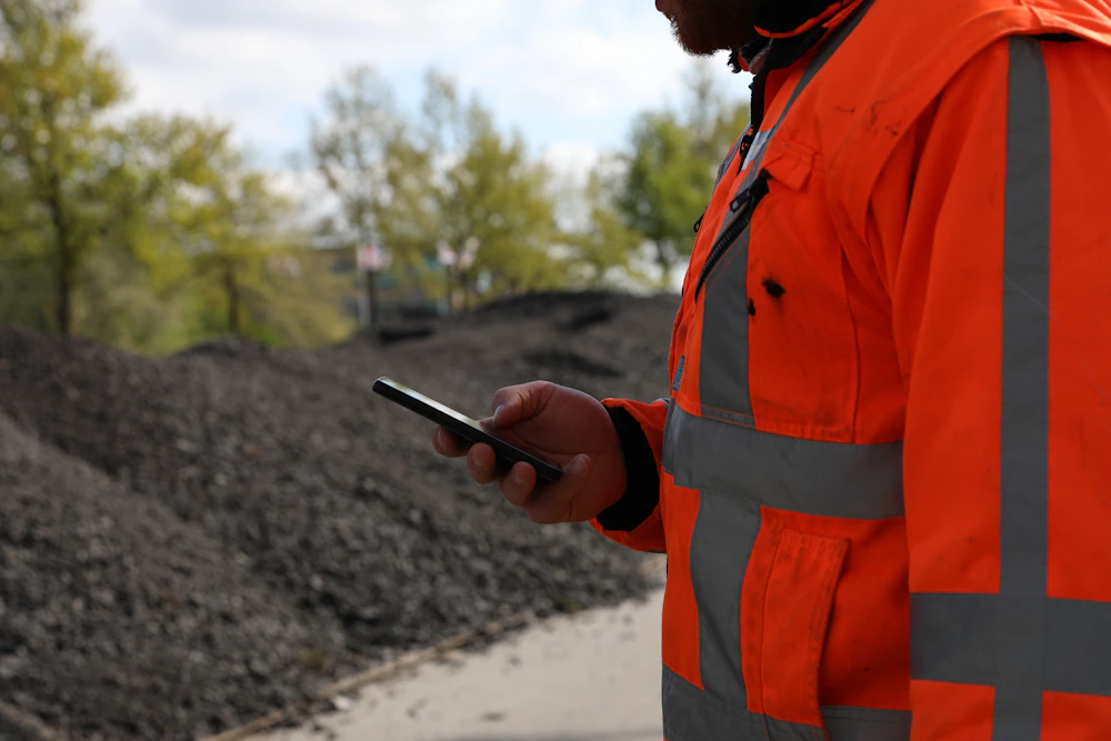 Mannelijke medewerker in reflecterende oranje jas kijkt op de telefoon in zijn hand waar hij de financiële administratie kan zien. Op de achtergrond liggen drie hopen met bouwmateriaal.