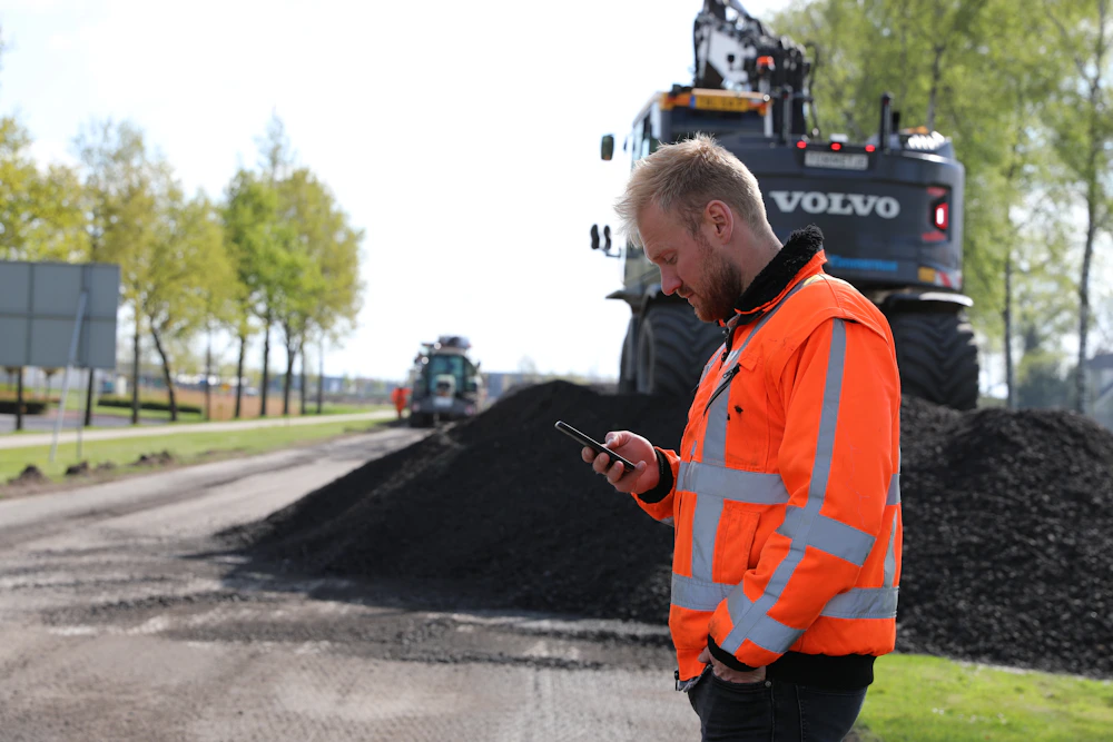Mannelijke bouwmedewerker bij een graafmachine die op zijn telefoon kijkt voor de digitale urenregistratie.