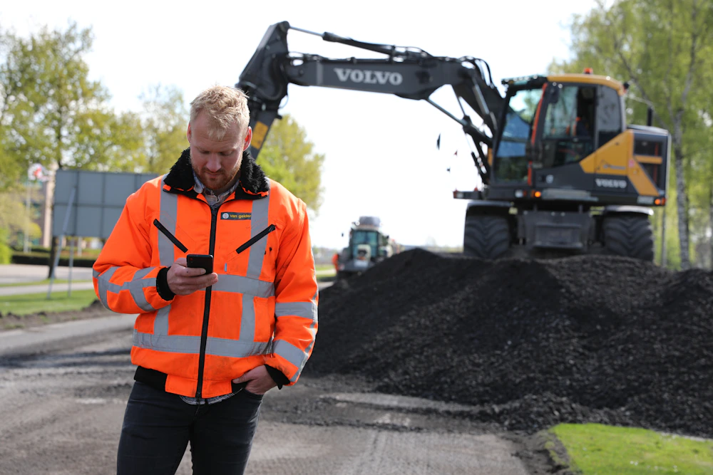 Een persoon in een fel oranje veiligheidsjas met reflecterende stroken kijkt op een telefoon; op de achtergrond staat een Volvo graafmachine op een hoop grind.