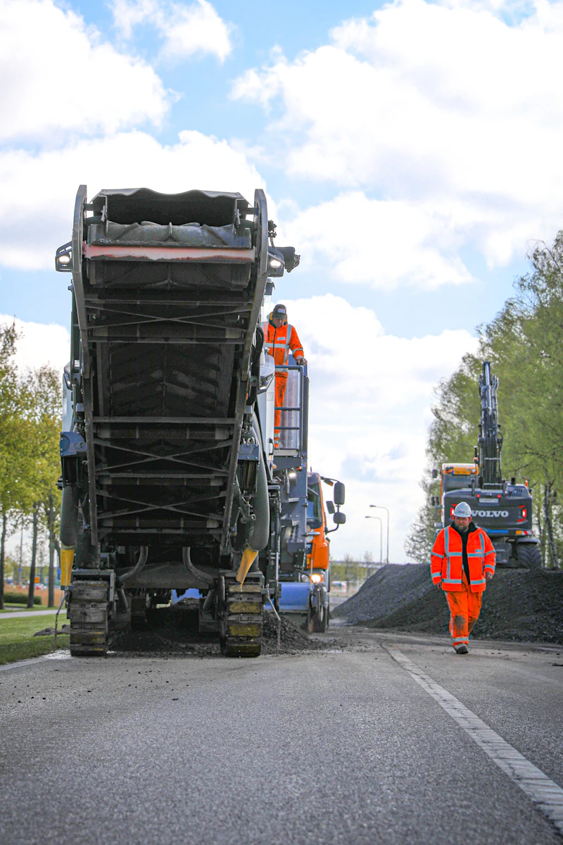 Arbeiders in oranje veiligheidskleding werken aan een weg met een grote asfaltpaver; een persoon staat op de machine, een ander loopt langs de weg.