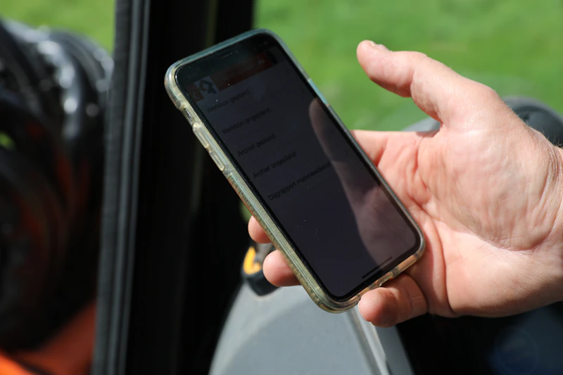 Een hand houdt een smartphone met een doorzichtige hoes vast; het scherm toont een lijst met opties, terwijl de groene buitenomgeving op de achtergrond vaag is.