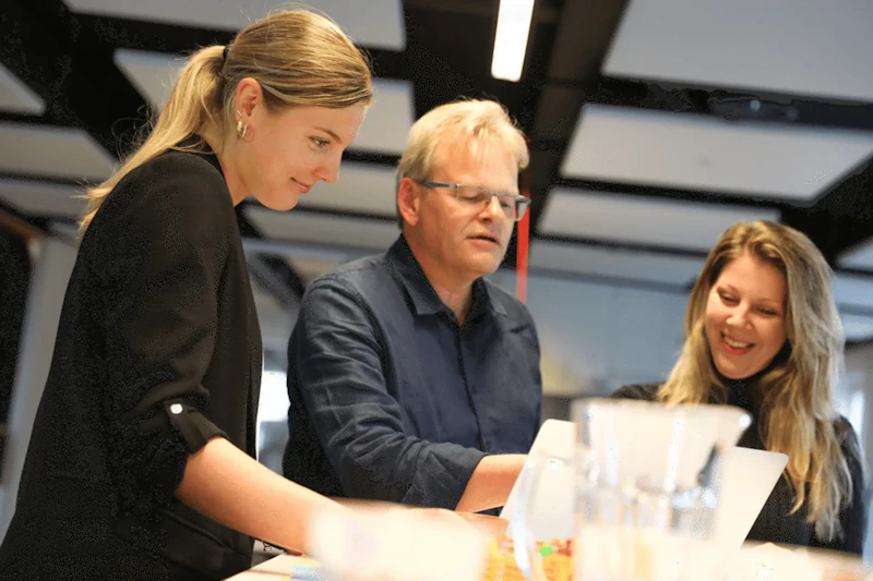 Drie personen staan rond een tafel met laptops en waterflessen, terwijl ze samen aan een document werken en glimlachen.