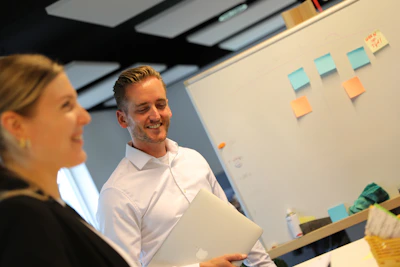 Twee personen lachen in een kantoor, met een whiteboard waarop blauwe en oranje post-its hangen; iemand houdt een MacBook vast.Werken bij VanMeijel