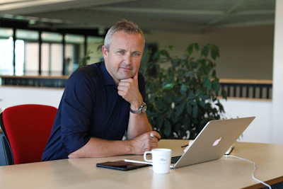 Een persoon zit aan een kantoorbureau met een open laptop, een witte mok en een telefoon voor zich; op de achtergrond groene planten en ramen.Plan een afspraak