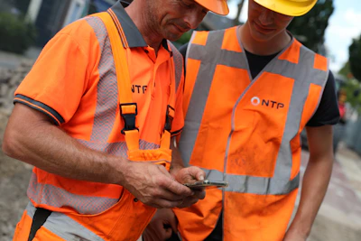 Twee personen in feloranje veiligheidsvesten met reflecterende stroken en helmen bekijken samen een smartphone op een bouwplaats.Papierloos werken