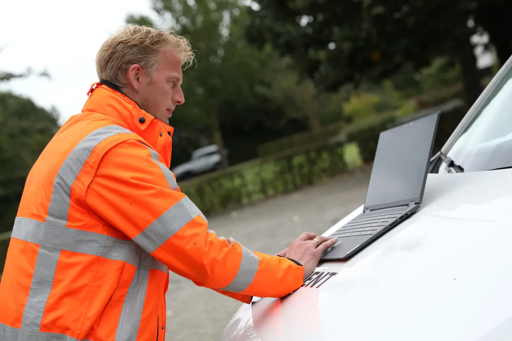 Een persoon in een fel oranje veiligheidsjas met reflecterende strepen gebruikt een laptop op de motorkap van een witte auto, buiten tussen bomen.