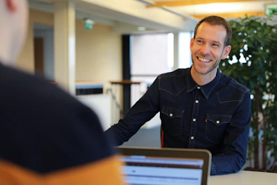 Een glimlachende persoon in een donkerblauw jeanshemd zit aan een tafel en praat met een andere persoon; op tafel ligt een laptop en op de achtergrond staan groene planten.Jouw partner voor het digitaliseren van al je bedrijfsprocessen