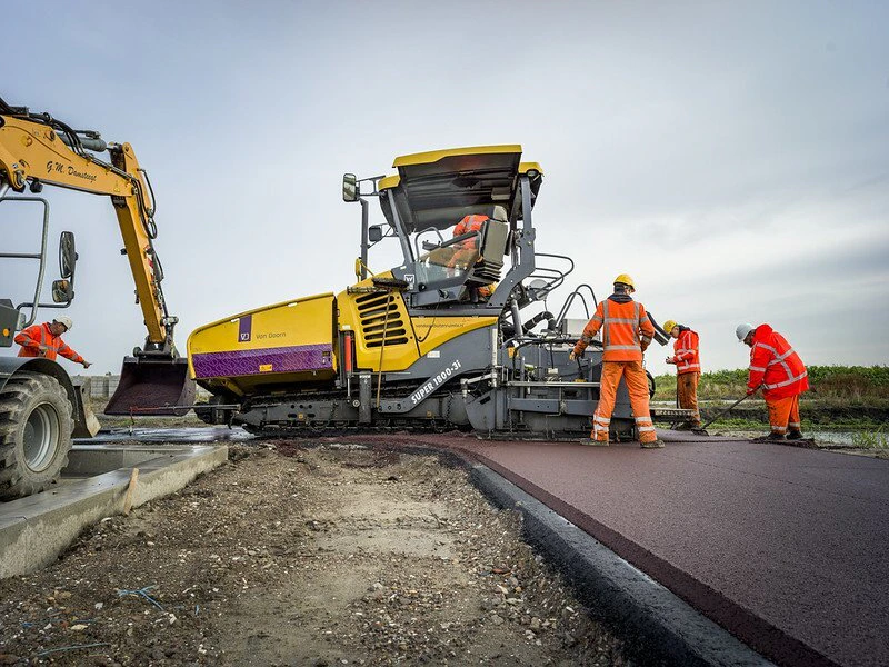 Arbeiders in oranje veiligheidsvesten en helmen leggen vers asfalt; een gele asfaltpaver werkt terwijl links een graafmachine staat.