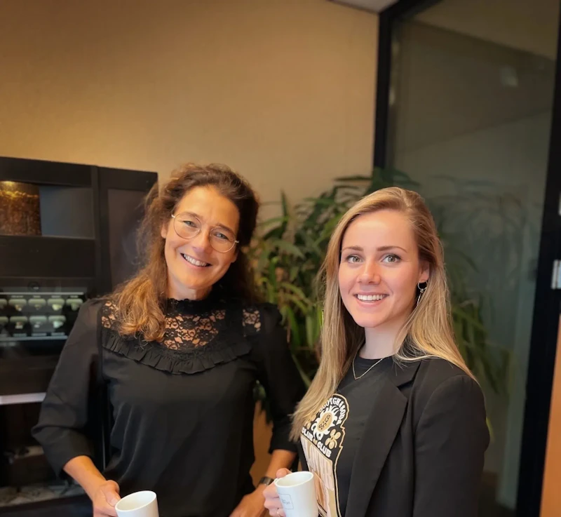 Twee personen staan in een kantoorruimte, glimlachen en houden witte koffiemokken vast; links met bril en kanten top, rechts in blazer met print, groene planten op de achtergrond.