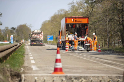 Werkauto op de web met daarvoor een oranje vrachtwagen waar drie bouwmedewerkers in oranje kleding aan het werk zijn. Op de voorgrond staat nog een pion.Snelle facturatie door digitale bouwplaatsregistraties