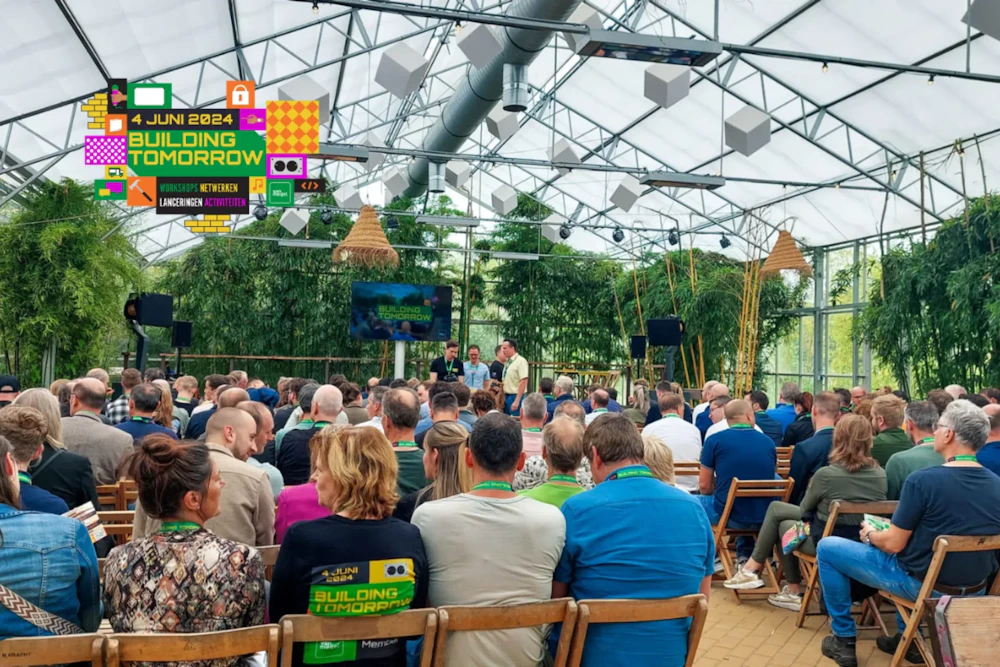 Publiek zit op houten stoelen in een glazen serre, gericht op een podium met scherm; banner Building Tomorrow en groene planten rondom.