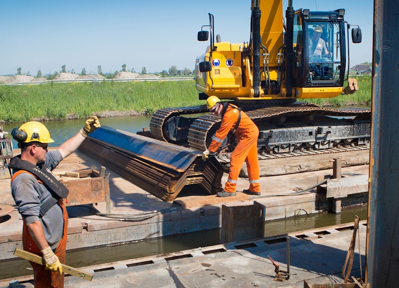 Twee mannen in werkkleding op een bouwplaats aan het water. Op de achtergrond is een gele kraan te zien.