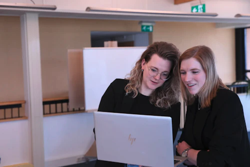 Twee personen bekijken samen een open HP-laptop in een kantoorruimte, staande naast een witte scheidingswand.