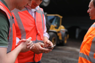 Drie personen in oranje veiligheidsvesten staan samen; één houdt een telefoon vast terwijl de anderen meekijken, met een gele heftruck op de achtergrond.83% van onze klanten in de burgerlijke en utiliteitsbouw beveelt ons aan!