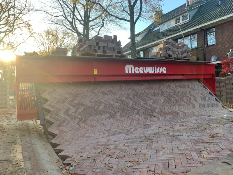 Twee arbeiders met helmen op een rode Meuwisse vrachtwagen vol stenen; een helling van metselstenen loopt uit de wagen, met herfstbladeren op de grond.