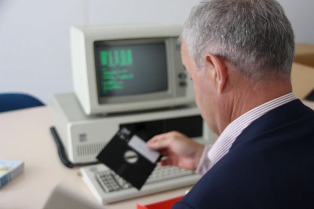 Een persoon met kort grijs haar in een donkerblauw pak houdt een floppy disk voor een oude computer met CRT-monitor en groen op zwart scherm.