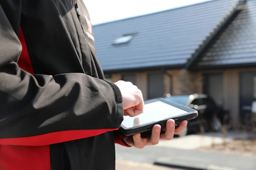 Een persoon in een zwarte jas met rode accenten houdt een tablet vast en tikt met een vinger op het scherm, buiten met een huis en auto op de achtergrond.