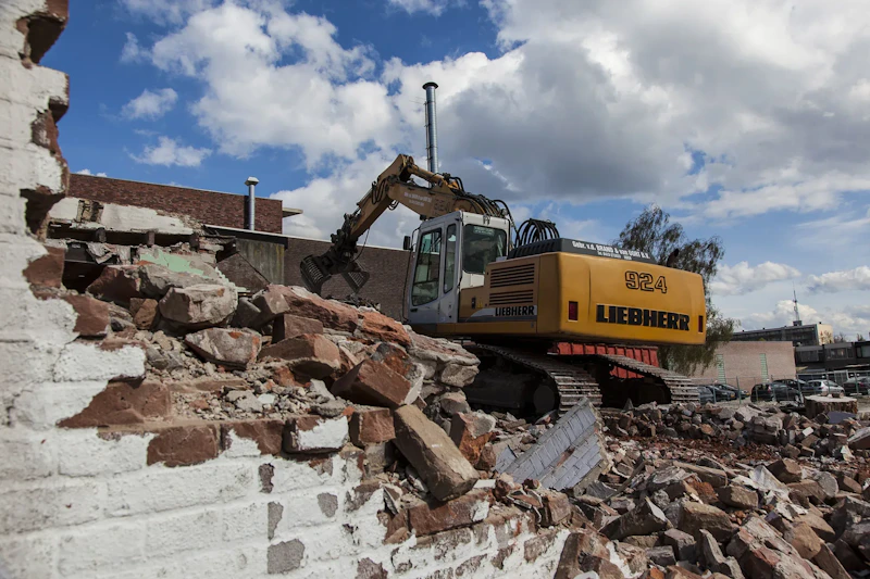 Gele Liebherr 924 graafmachine staat op puin van een afgebroken muur, met stenen en gebouwen op de achtergrond onder een blauwe hemel.