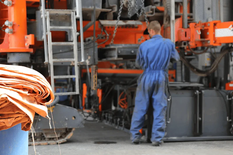 Een persoon in een blauwe overall staat voor een grote industriële machine met een oranje frame en kettingen in een drukke werkplaats.
