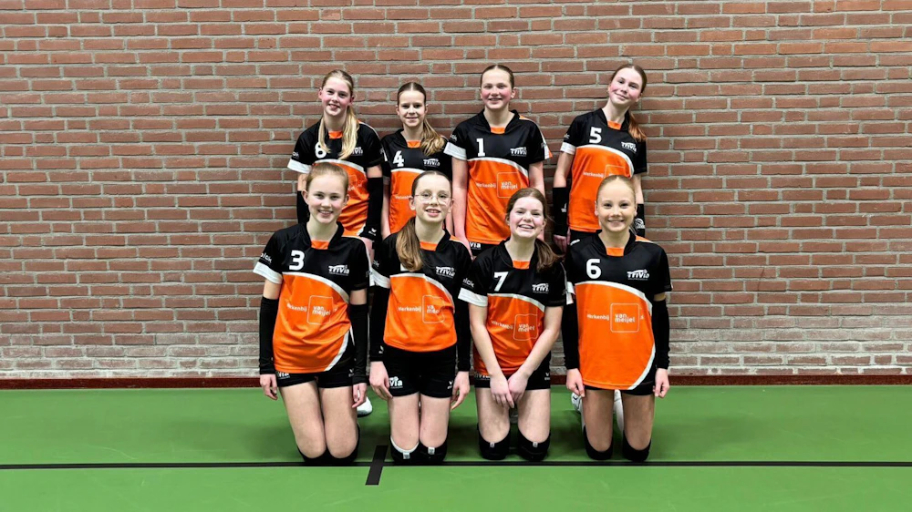 acht jonge volleybalspelers in oranje-zwarte uniformen poseren voor een teamfoto tegen een bakstenen muur op een groene zaalvloer