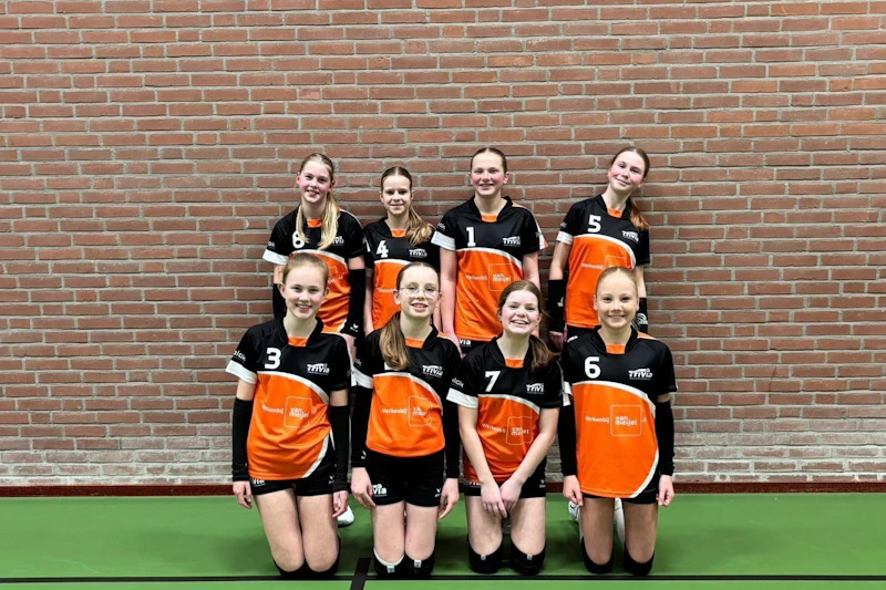 Acht spelers in oranje-zwart volleybaluniformen staan en knielen voor een bakstenen muur in een sporthal, lachend naar de camera.