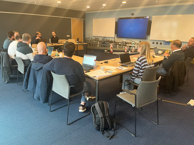 Een groep mensen zit rondom een lange vergadertafel met laptops en notities; aan de muur een groot scherm toont een blauw beeld.