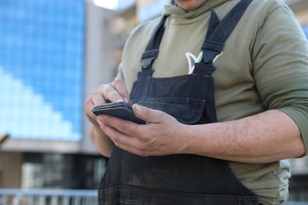 Persoon in olijfgroene hoodie en donkerblauwe overalls houdt met beide handen een smartphone vast; achtergrond een blauw glazen gebouw, vaag.