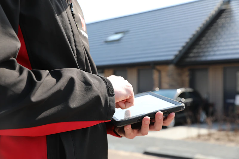 Een persoon in een zwarte jas met rode voering houdt buiten een tablet vast; achtergrond met een huis en geparkeerde auto.