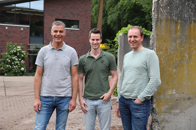 Drie personen staan buiten voor een bakstenen gebouw met groen, glimlachen naar de camera; casual kleding en jeans.