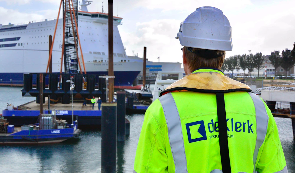 Persoon in fluorgeel veiligheidsvest en witte helm staat op een havenkade, met een groot cruiseschip en kranen op de achtergrond.