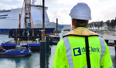 Persoon in fluorgeel veiligheidsvest en witte helm staat op een havenkade, met een groot cruiseschip en kranen op de achtergrond.Inzicht en grip op projecten