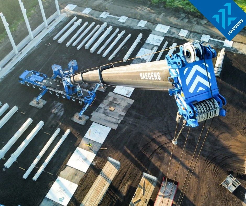 Blauwe hijskraan (HAEGENS) tilt een lange buis op een bouwplaats; rondom liggen witte stalen pijpen en bouwmaterialen op een donkere ondergrond.