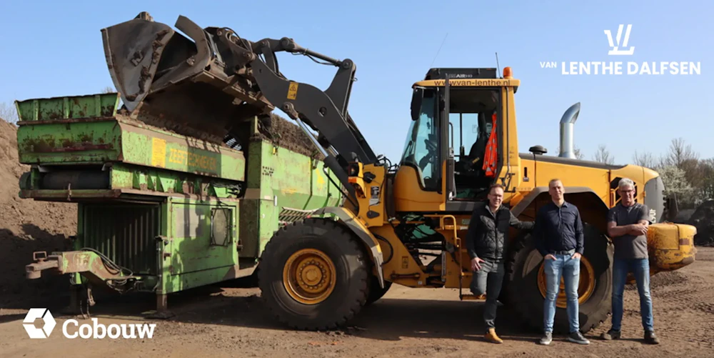 Drie personen staan naast een grote gele wiellader die materiaal in een groene machine leegt op een bouwterrein onder een blauwe lucht.