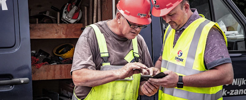 Twee personen met rode veiligheidshelmen en fluorescerende veiligheidsvesten bekijken samen een smartphone bij een gereedschapswagen.