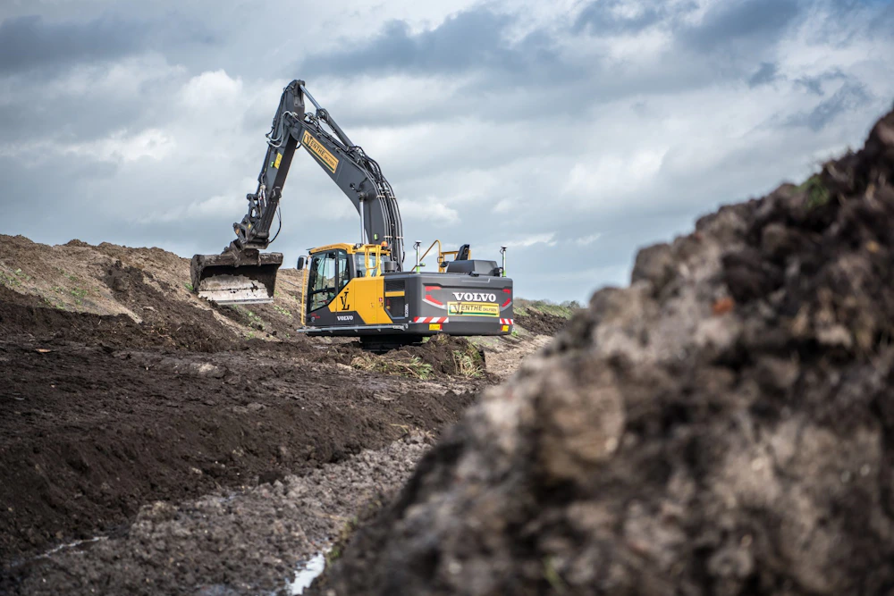 Volvo graafmachine op een bouwterrein, hij graaft in een diepe geul tussen modder en grond, onder een bewolkte hemel.