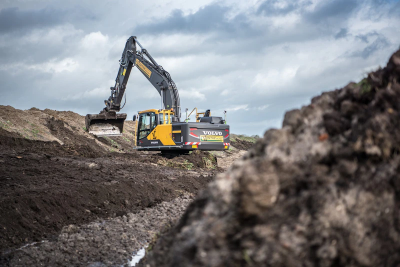 Volvo graafmachine op een bouwterrein, hij graaft in een diepe geul tussen modder en grond, onder een bewolkte hemel.