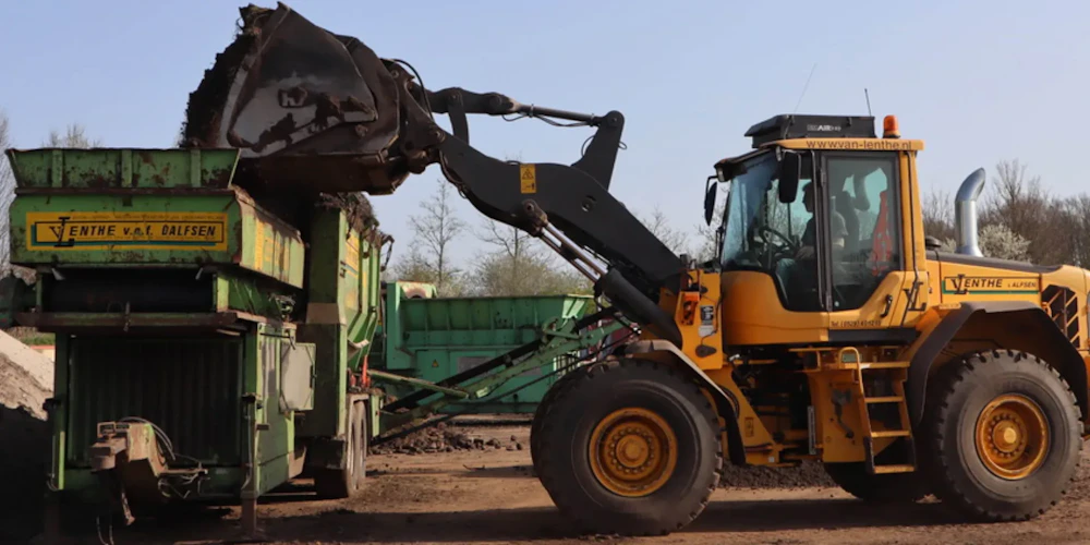 Een gele wiellader laadt aarde in een groene kipperwagen op een bouwplaats; bomen staan achter de machines.