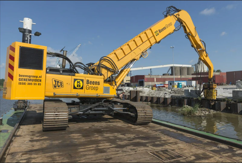 Gele hijskraan op een drijvend werkplatform bij een haven, met lange uitschuifbare arm en bedrijfslogo’s Beens Groep en JCB, tegen een blauwe lucht.