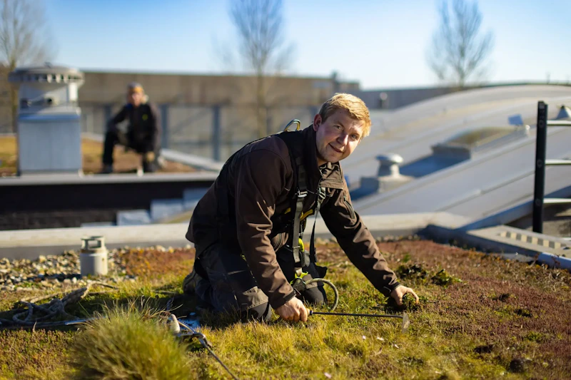 Een persoon knielt op een groendak met veiligheidsuitrusting en harnas, bezig met gereedschap; op de achtergrond zit iemand.