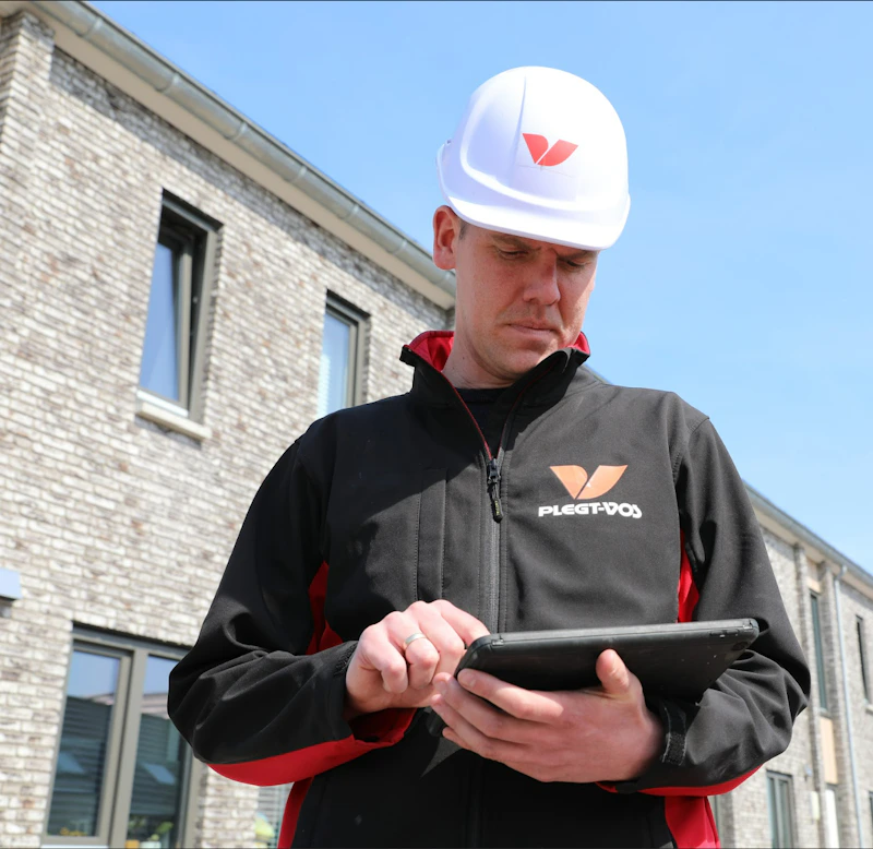 Een persoon met witte veiligheidshelm en een zwarte jas met oranje V-logo staat buiten bij een stenen gebouw en bekijkt een tablet.