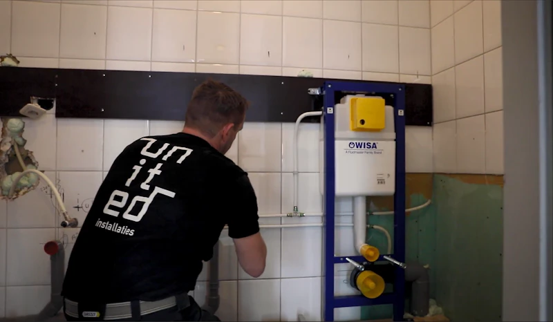 Een technicus werkt aan een wandcloset-inbouwframe tegen een betegelde muur; wit reservoir in een blauwe metalen omlijsting met gele dopjes.