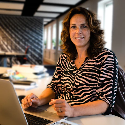 Een persoon met golvende bruine haren zit aan een bureau met een laptop, glimlacht en draagt een zebra-print blouse in een moderne kantoorruimte.Auteur blog