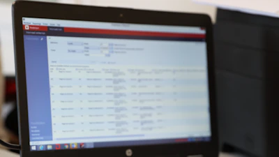 Een laptop met een onscherp scherm waarop een softwarevenster verschijnt: linkerpaneel blauw en een tabelachtig hoofdvenster met rijen en kolommen.