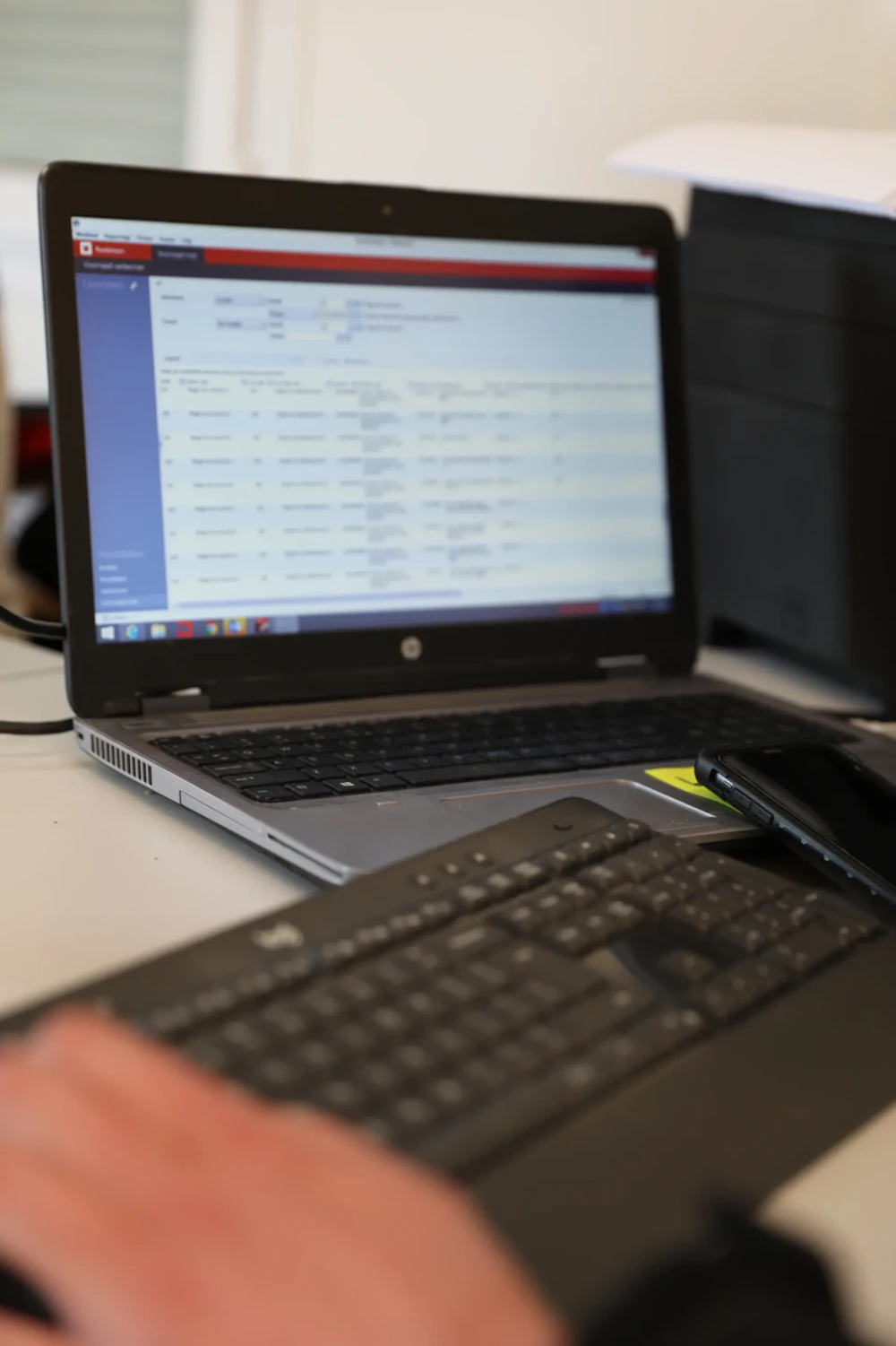 Open laptop op een bureau met een wazig scherm; voorgrond een tweede toetsenbord en een smartphone, met een hand op het toetsenbord.