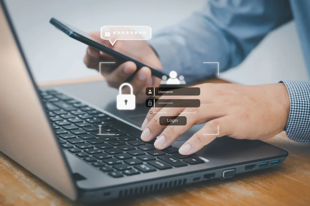 Een persoon typt op een laptop terwijl een smartphone vastgehouden wordt; overlay toont een beveiligde inlog met gebruikersnaam, wachtwoord en login-knop.