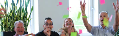 Vier mensen vieren in een kantoor, armen omhoog en lachend, met kleurrijke Post-its in de lucht en een groene plant op de achtergrond.Bedankt voor je aanvraag