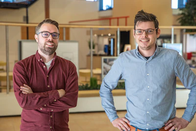 Twee personen met brillen staan in een open kantoorruimte en glimlachen; links rood overhemd, armen over elkaar, rechts lichtblauw overhemd met hand op de heup.Onze visie bij VanMeijel